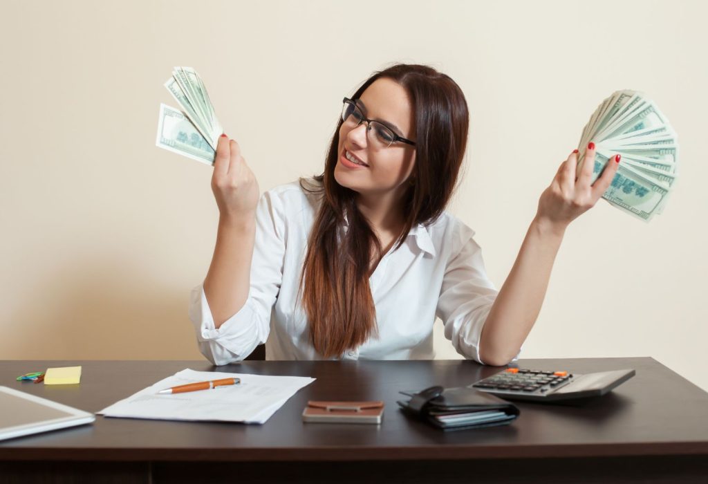 Smiling Bookkeeper girl sitting at her desk holding money. Representing Financial Rewards for Bookkeepers.